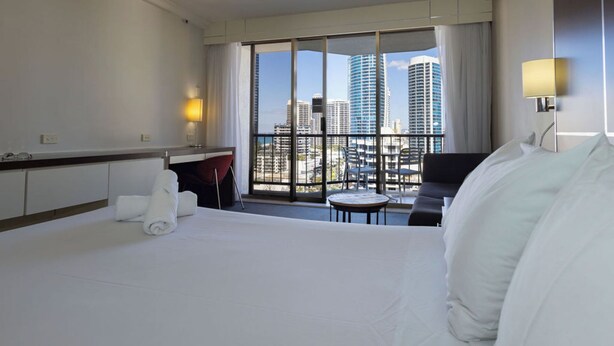 Ein Hotelzimmer mit Bett, Schreibtisch, Stuhl und Blick auf die Stadt durch große Fenster.