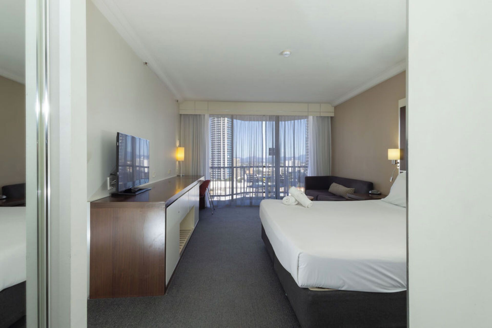 Une chambre d’hôtel avec un lit, un bureau doté d’une télévision, une chaise et une vue sur la ville grâce à de grandes fenêtres.