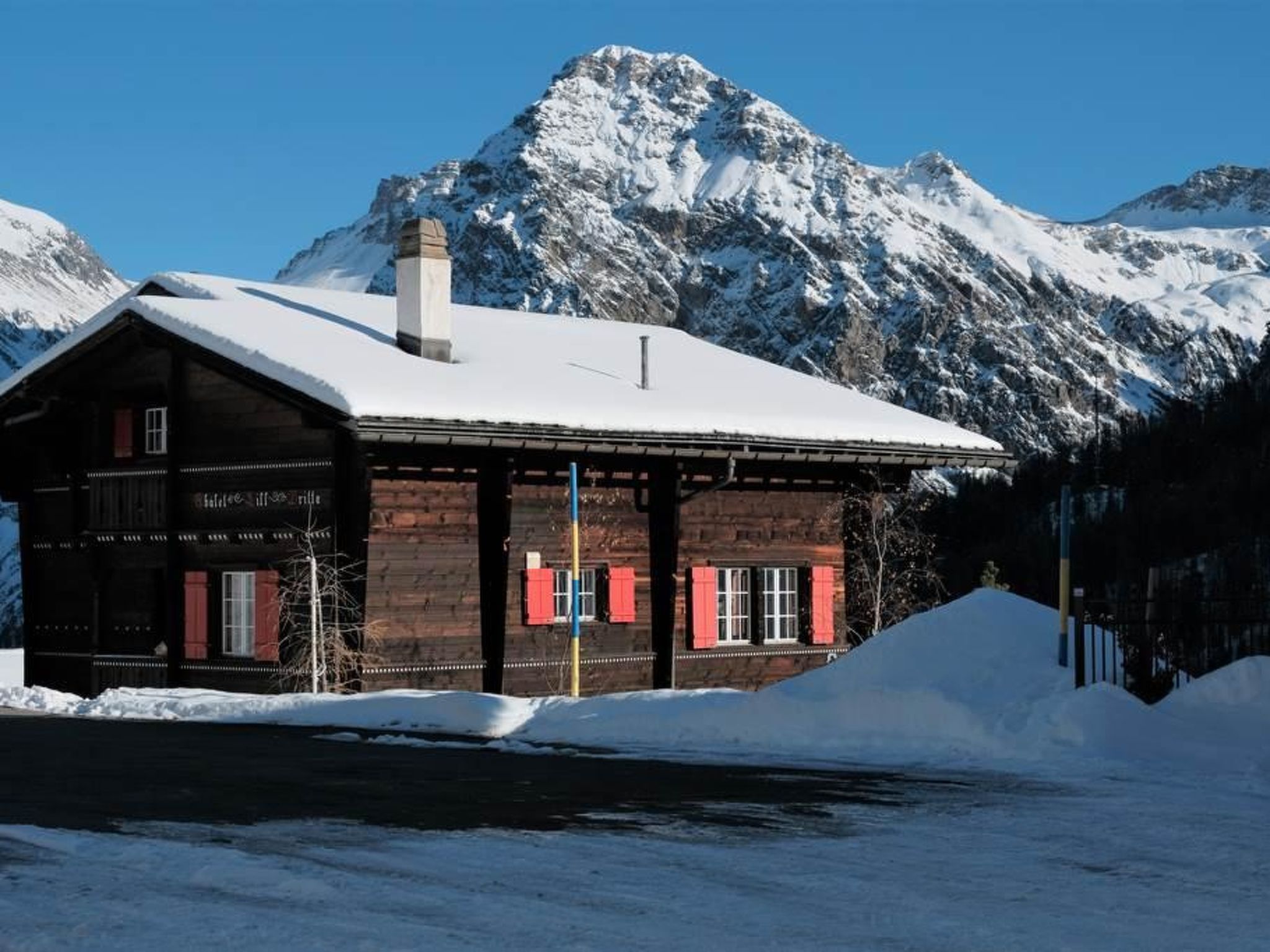 Un edificio de madera con techo nevado, una chimenea y contraventanas rojas, con montañas nevadas al fondo.