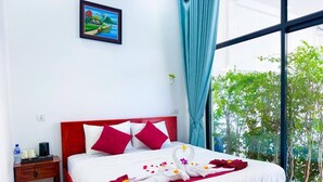 A hotel room with a bed, a nightstand, a painting on the wall, and a view of greenery outside.