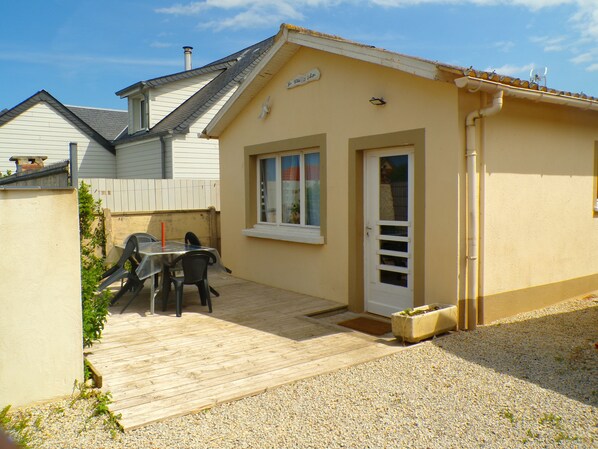 A small, single-story building with a wooden deck, a table with chairs, and a door with a window.