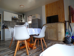 A compact kitchen with modern appliances, a dining area with white chairs, and a television mounted on a wooden panel.