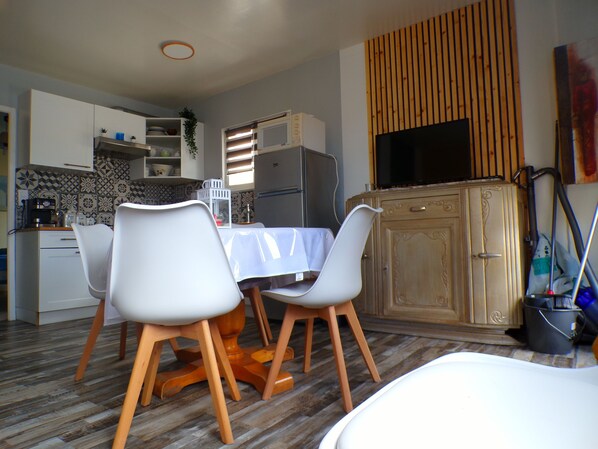 A compact kitchen with modern appliances, a dining area with white chairs, and a television mounted on a wooden panel.