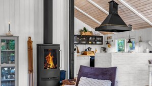 Una habitación acogedora con chimenea de leña, un sillón cómodo y una zona de cocina con armarios blancos y una lámpara colgante.