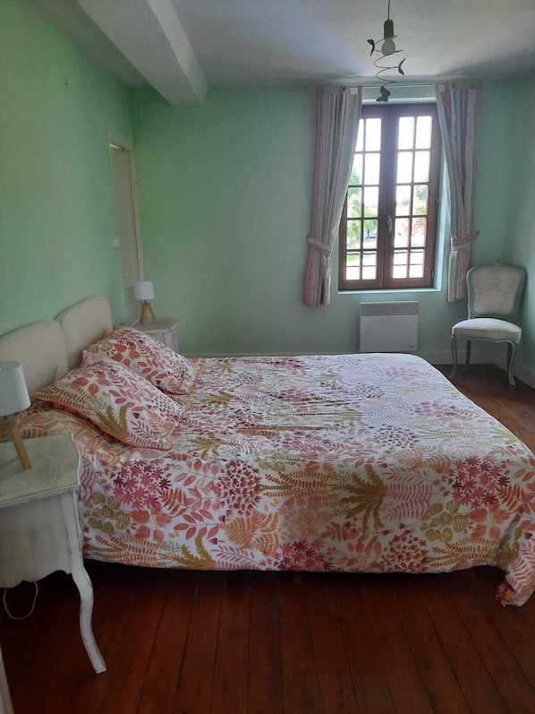 A bedroom with a large bed, a bedside table with a lamp, a chair, a window with curtains, and a radiator.