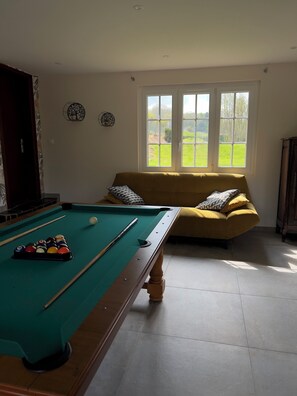A pool table with balls and cues, a sofa with cushions, and a window with a view of greenery.