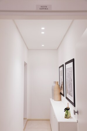 Un pasillo con pared blanca, techo con iluminación empotrada y un armario blanco con dos cuadros enmarcados y un jarrón decorativo.