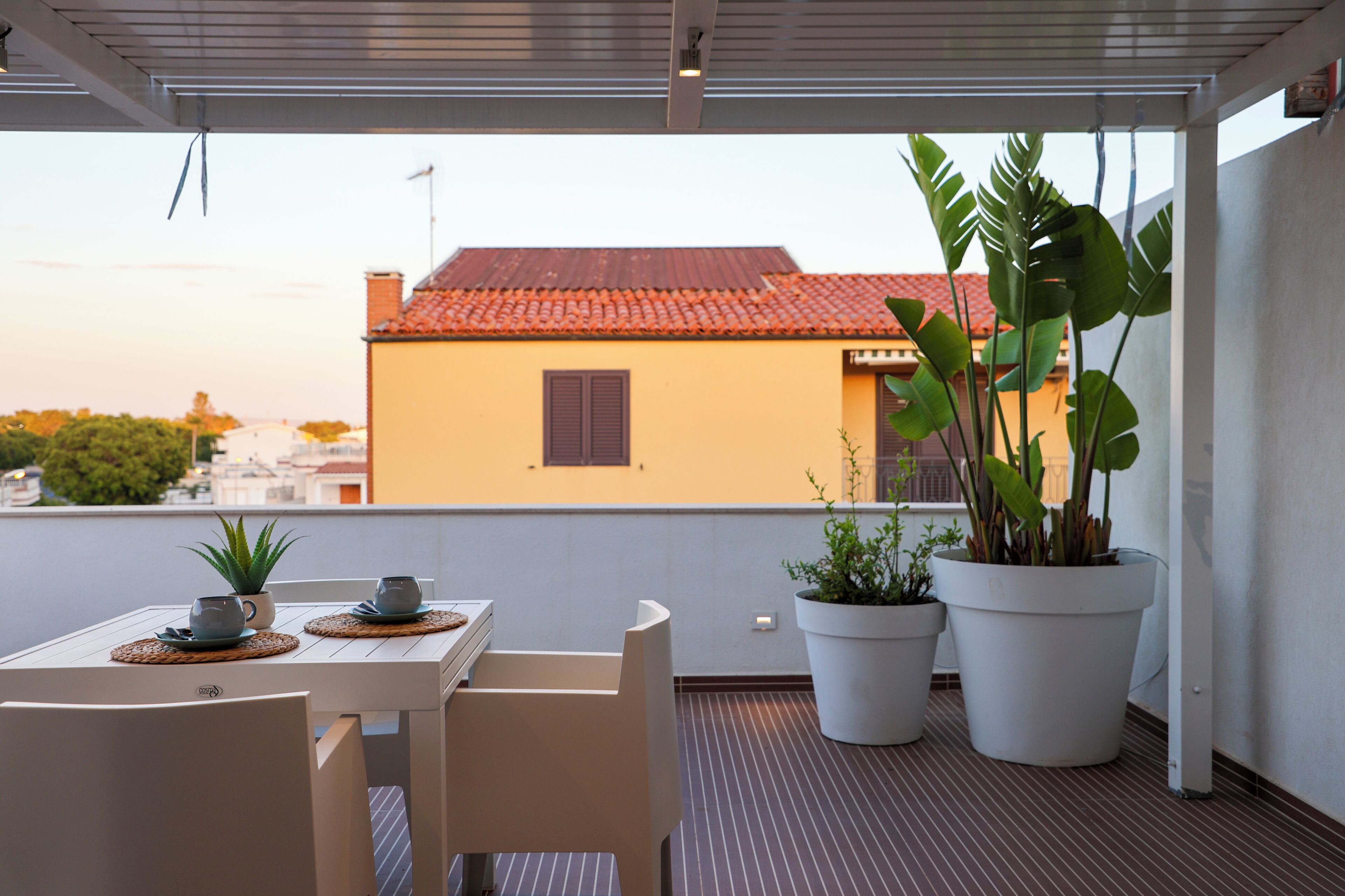 Ein Balkon mit Tisch für Zwei, Topfpflanzen und Blick auf ein Gebäude mit roten Ziegeln.