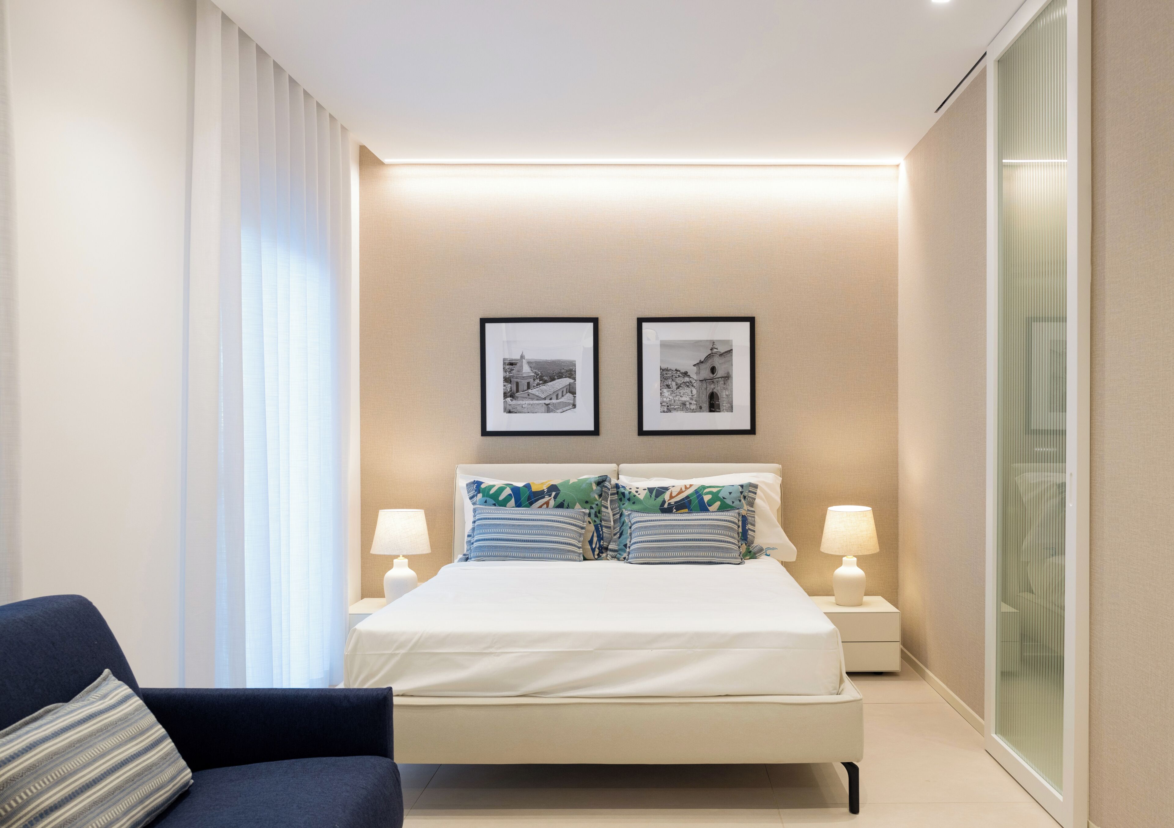 A modern hotel room with a large bed, two bedside tables with lamps, a nightstand, a chair, and two framed pictures on the wall.