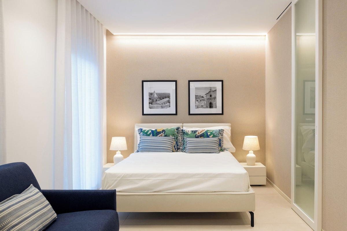 Quarto de hotel moderno com uma cama grande, duas mesas de cabeceira com luminárias, um criado-mudo, uma cadeira e duas fotos emolduradas na parede.