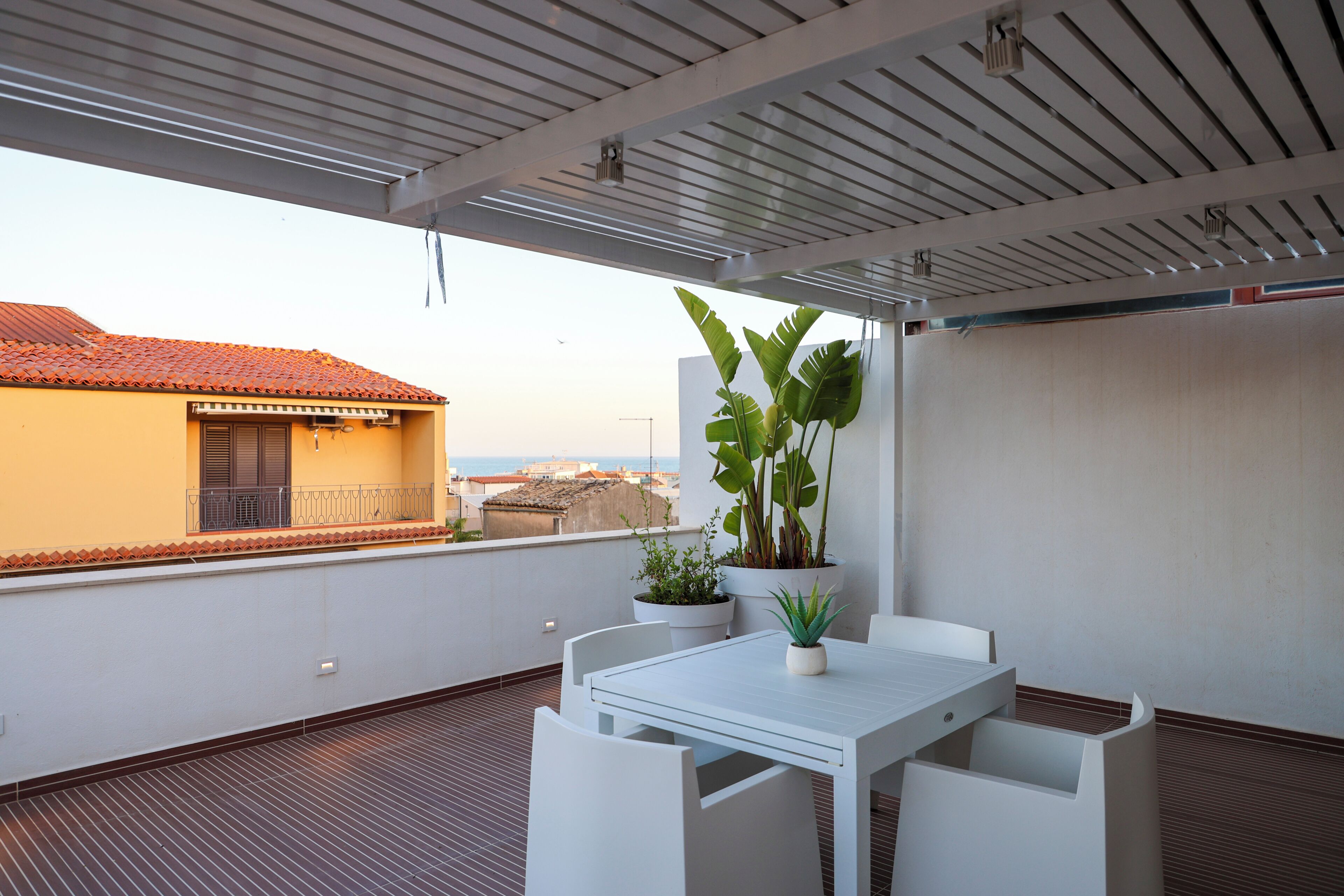 Eine Dachterrasse mit weißem Tisch und Stühlen, Topfpflanzen und Blick aufs Meer.