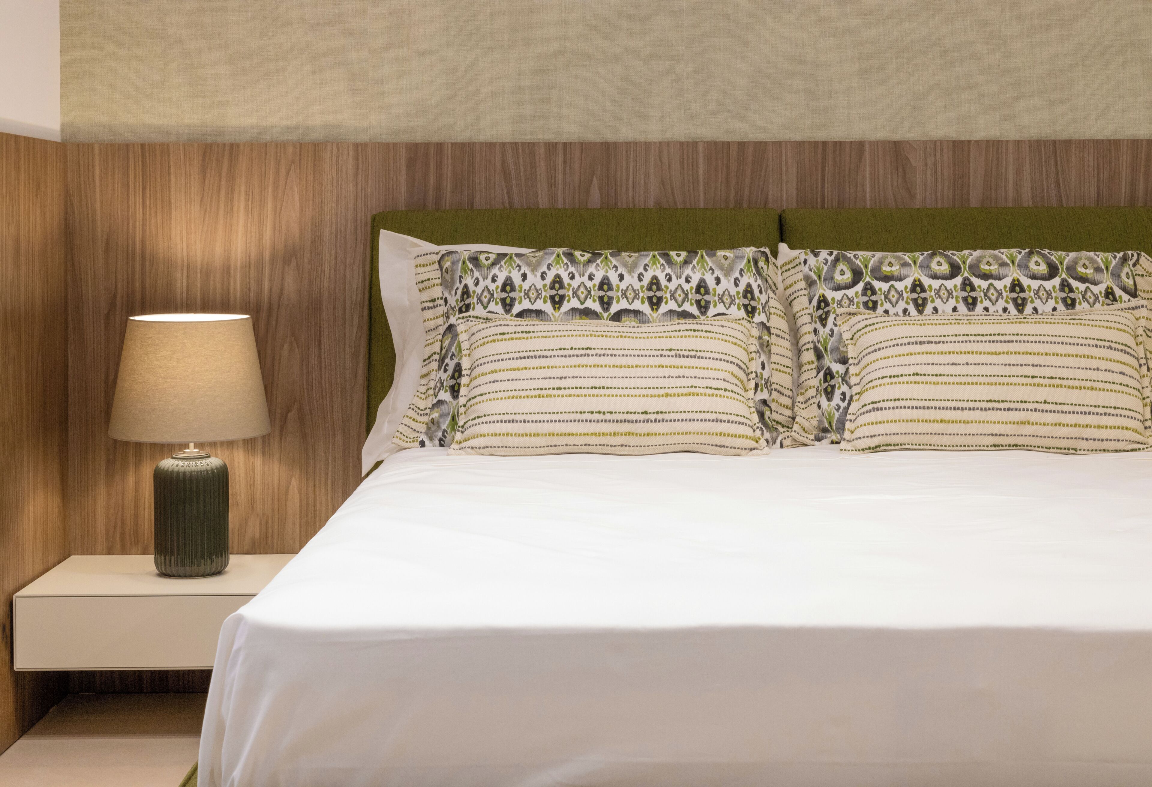 Una cama bien tendida con un cabecero verde, una lámpara de noche y almohadones estampados.