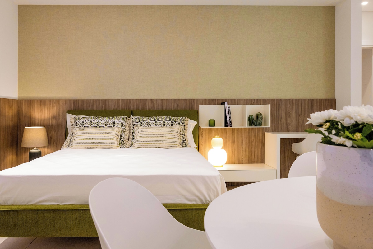 Quarto de hotel com cama, mesas de cabeceira, abajur, cadeira e um vaso com flores.