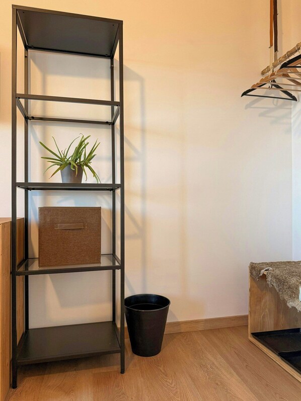 A room with a black metal shelving unit, a potted plant, a brown storage box, a black trash bin, and a wooden table with a cloth.