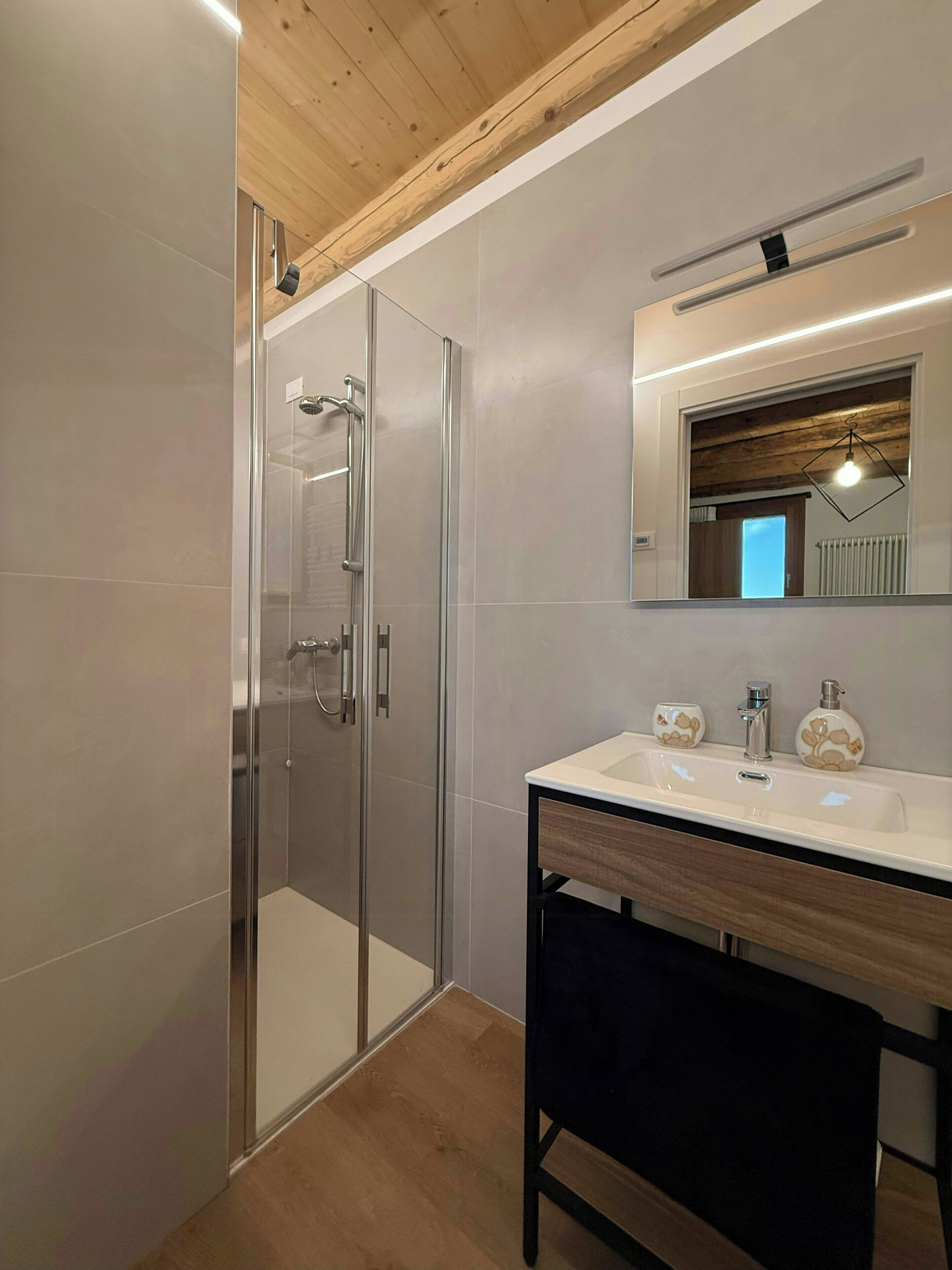 Un bagno moderno con doccia vetrata, soffitto in legno e un mobile lavabo con specchio e due erogatori di sapone.