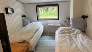 Um quarto pequeno e acolhedor com duas camas de solteiro, uma escrivaninha de madeira, uma janela com vista para o verde e uma televisão na parede.