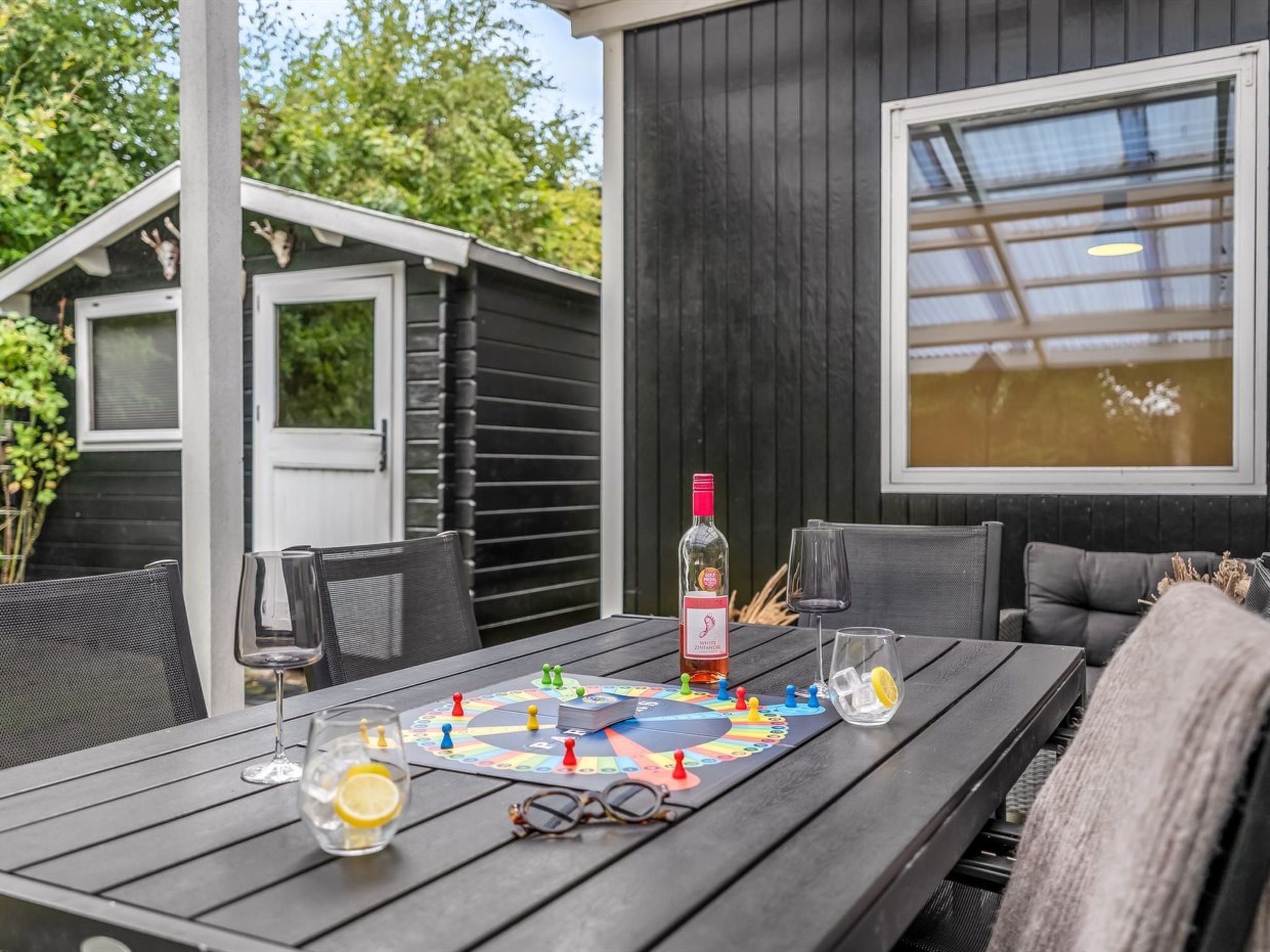 An outdoor dining area with a table set for two, featuring a board game, glasses, and a bottle of white wine.