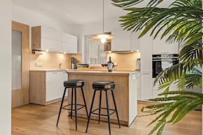 A modern kitchen with wooden cabinets, a central island with stools, a coffee machine, and a wine bottle. - Vacation home with a terrace hot tub next to the ski lift (Ruhpolding)