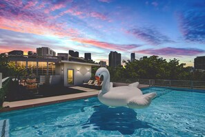 Un edificio moderno con facciata in vetro, una piscina con un galleggiante a forma di cigno e uno skyline urbano sullo sfondo.