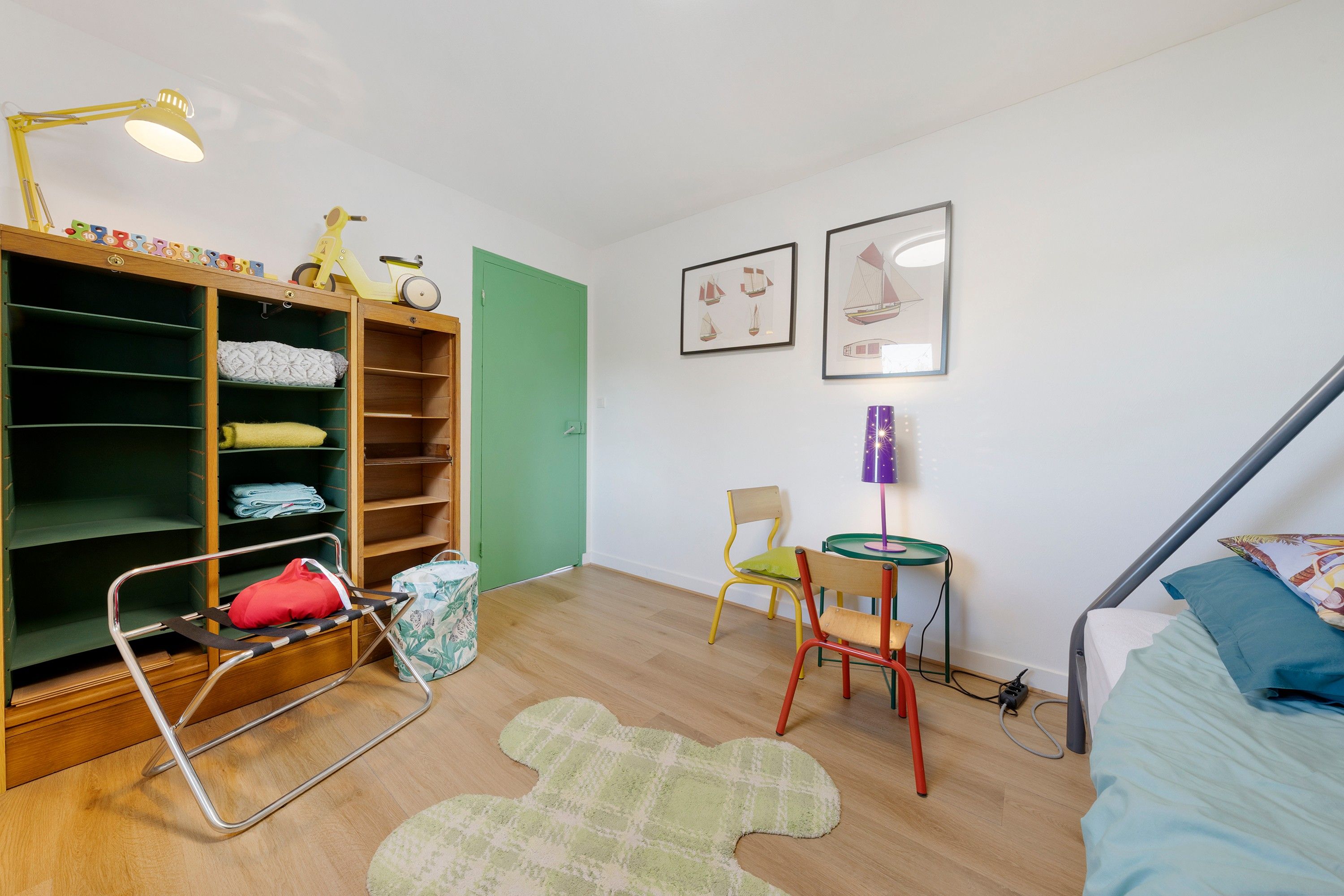 Een kinderkamer met een houten kast, een groene deur, een klein tafeltje met een paarse lamp, een groene stoel en een bed met een blauwe deken.