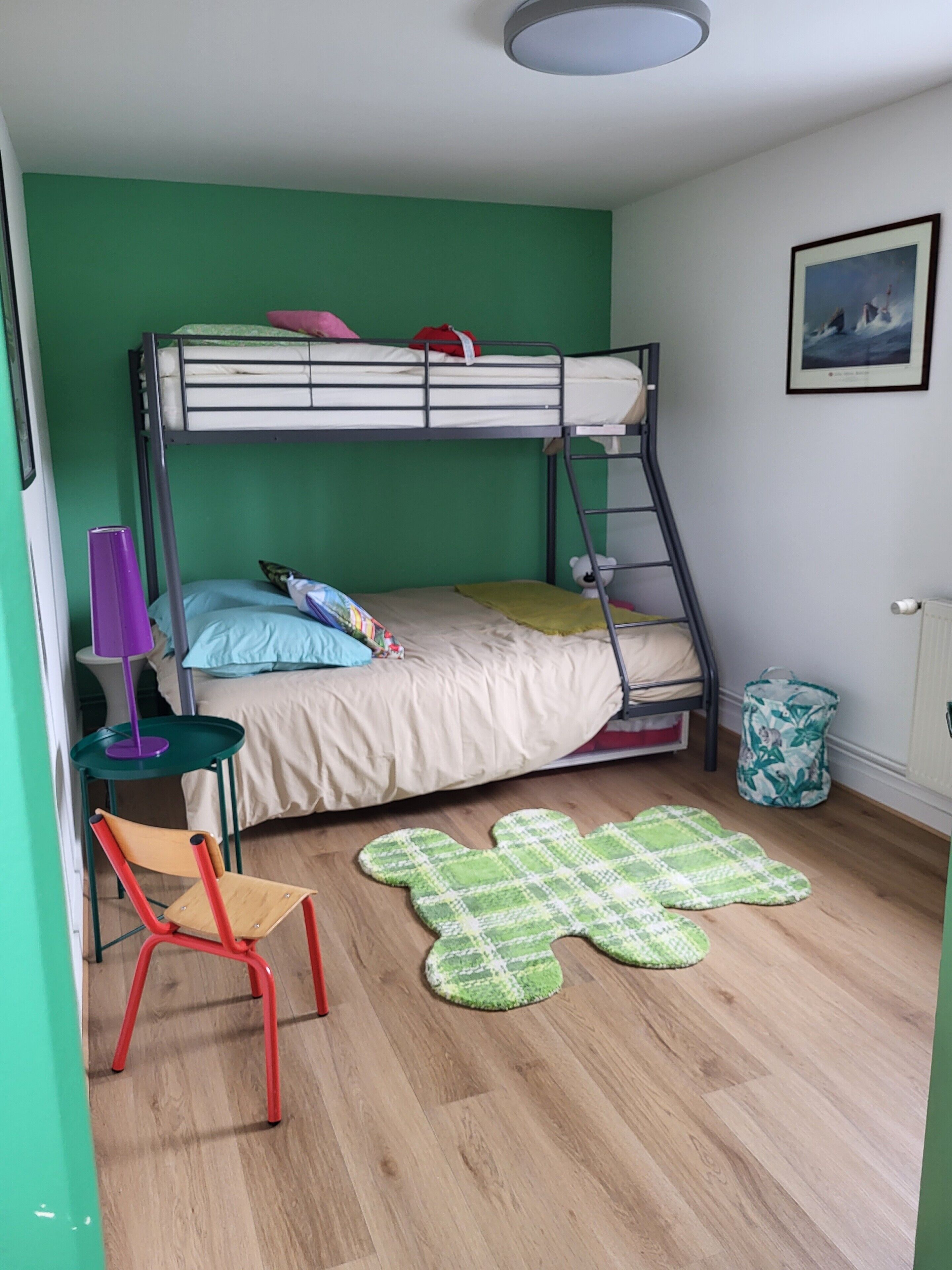 Une chambre avec un lit superposé, un mur vert, une petite table avec une lampe violette, une chaise rouge et un tapis à motifs verts.