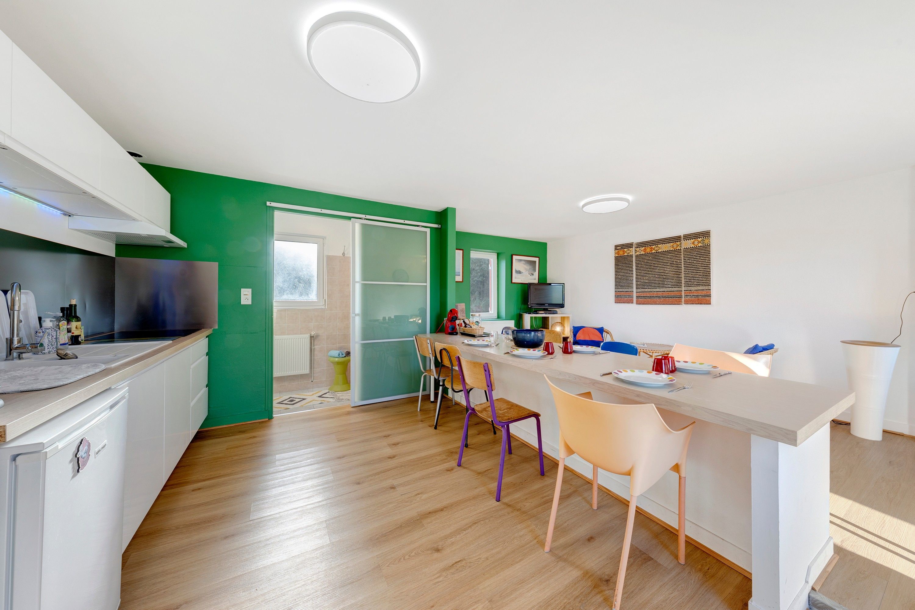 Eine moderne Küche mit grüner Akzentwand, Holzboden und einem Essbereich mit bunten Stühlen.