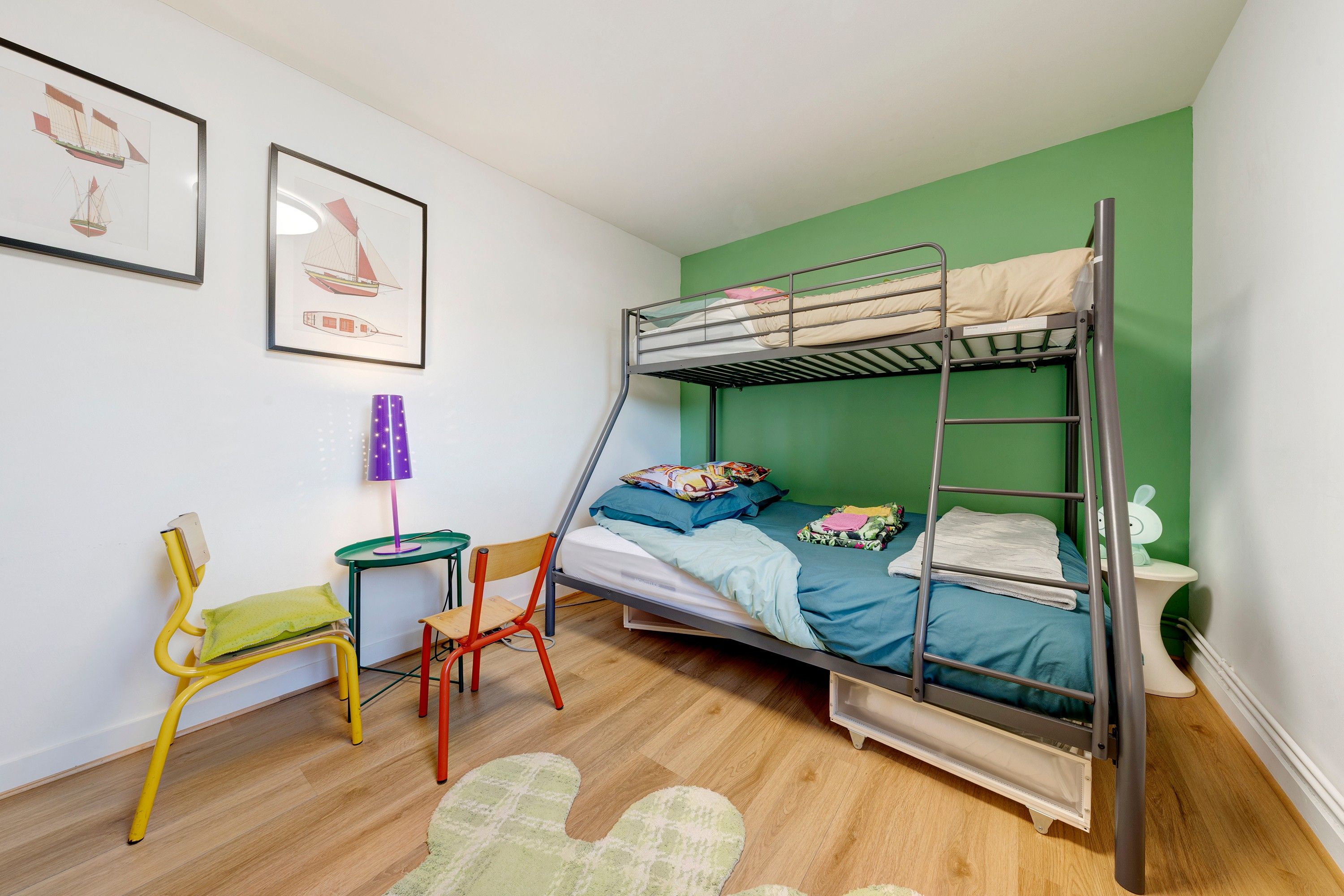 Une chambre avec un lit superposé, un mur vert, un bureau avec une chaise, une petite table avec une lampe et un tableau encadré accroché au mur.