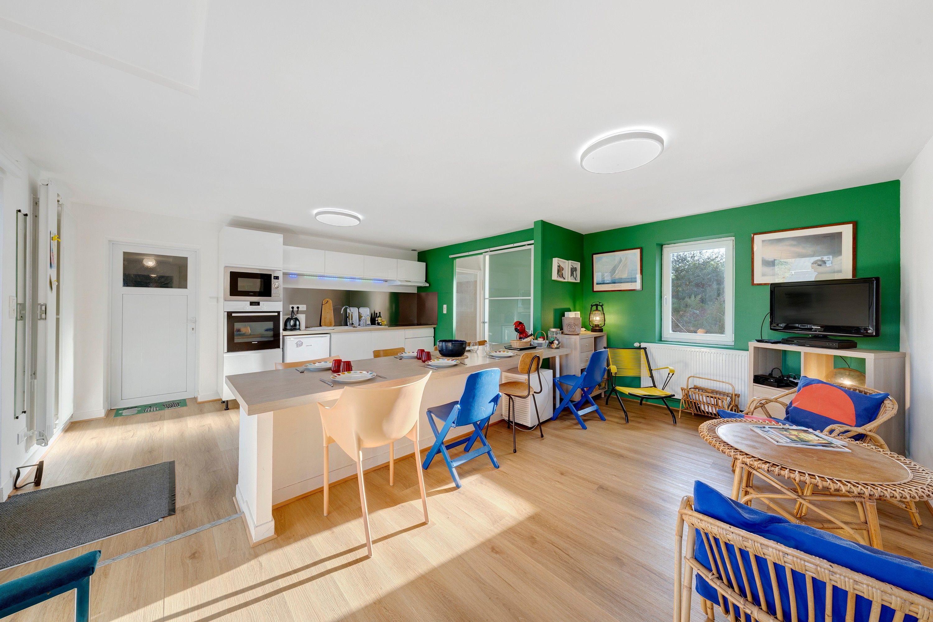 Een moderne keuken met witte kasten, een eethoek met blauwe en gele stoelen, een televisie en een groene accentmuur.
