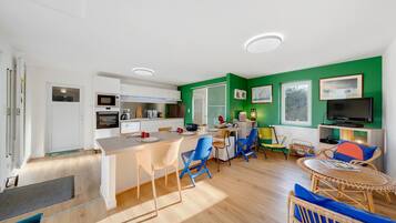 Een moderne keuken met witte kasten, een eethoek met blauwe en gele stoelen, een televisie en een groene accentmuur.
