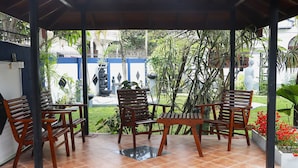 A covered outdoor seating area with wooden furniture and a view of a garden with statues and plants.