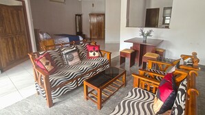 A living room with a sofa, armchairs, a coffee table, and a dining table with chairs. There is a wooden door and a window with blinds.