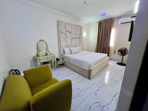 A hotel room with a bed, a vanity with a mirror, a yellow armchair, a standing fan, and a marble floor. - Centenary Hotel (Abuja)