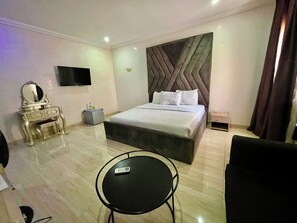 A hotel room with a bed, a vanity with a mirror, a flat-screen TV, a small table, and a chair. - Centenary Hotel (Abuja)