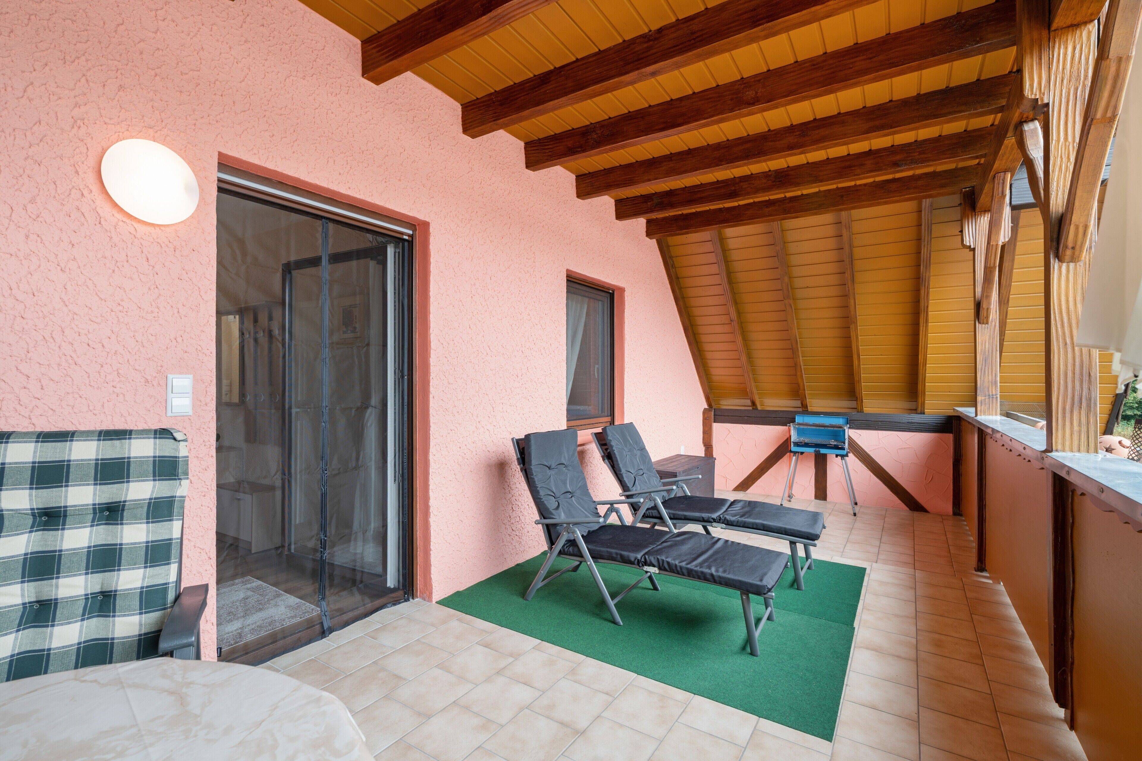 Eine sonnige Terrasse mit einer grünen Matte, zwei Liegestühlen, einem kleinen Tisch und einem blauen Grill.