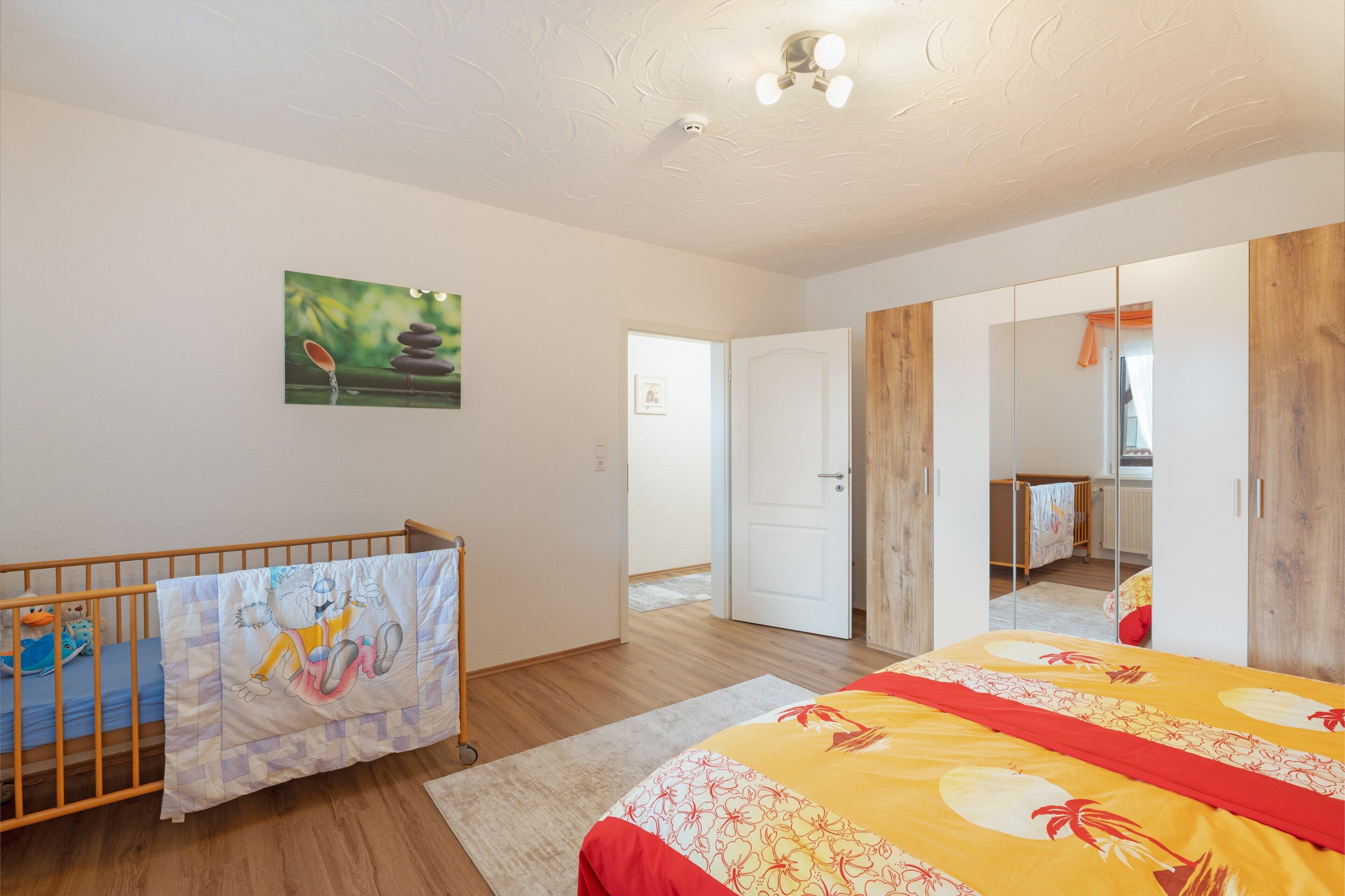 Ein Schlafzimmer mit einem Kinderbett, einem Bett mit floralem Bettzeug, einem Bild an der Wand und einem hölzernen Kleiderschrank.