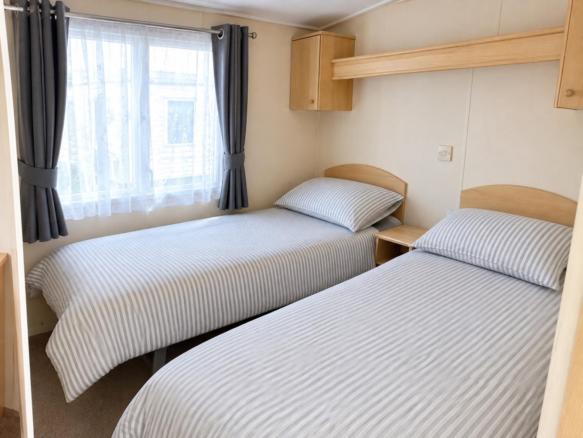 Una habitación con dos camas individuales, un cabecero de madera, una pequeña estantería y una ventana con cortinas.