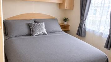 Una cama individual con cabecera, ropa de cama gris, una almohada decorativa, una mesita de noche con una planta y una ventana con cortinas grises.