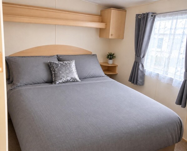 A single bed with a headboard, grey bedding, a decorative pillow, a small bedside table with a plant, and a window with grey curtains.