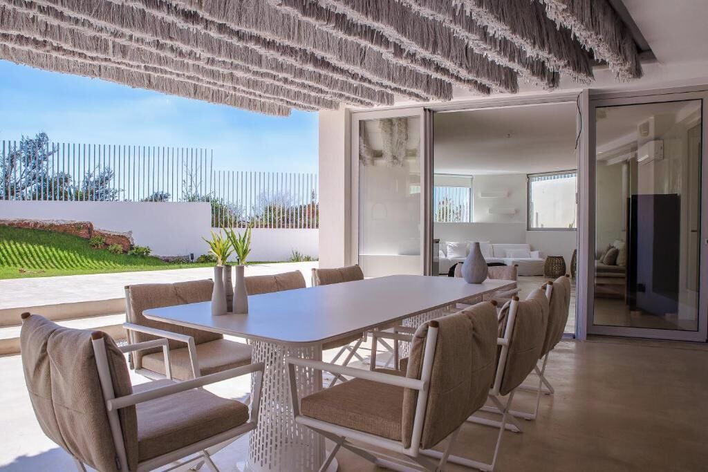 An outdoor dining area with a table and chairs, a glass door leading to a kitchen, and a view of a green landscape.
