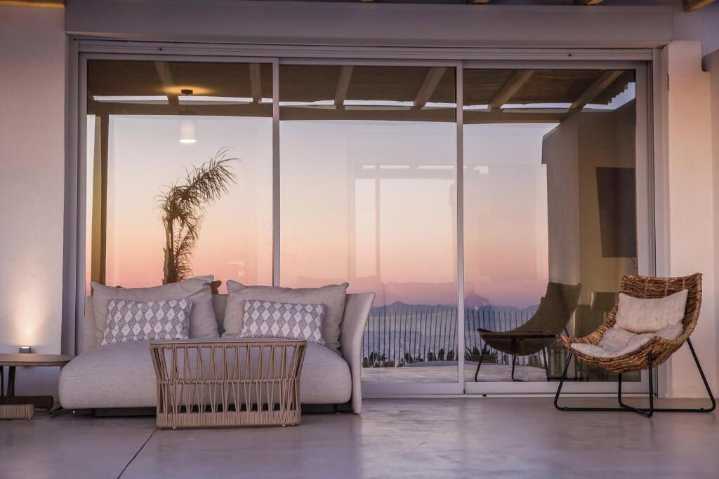 A modern living room with a sofa, a wicker chair, and a large sliding glass door leading to a balcony with a view.