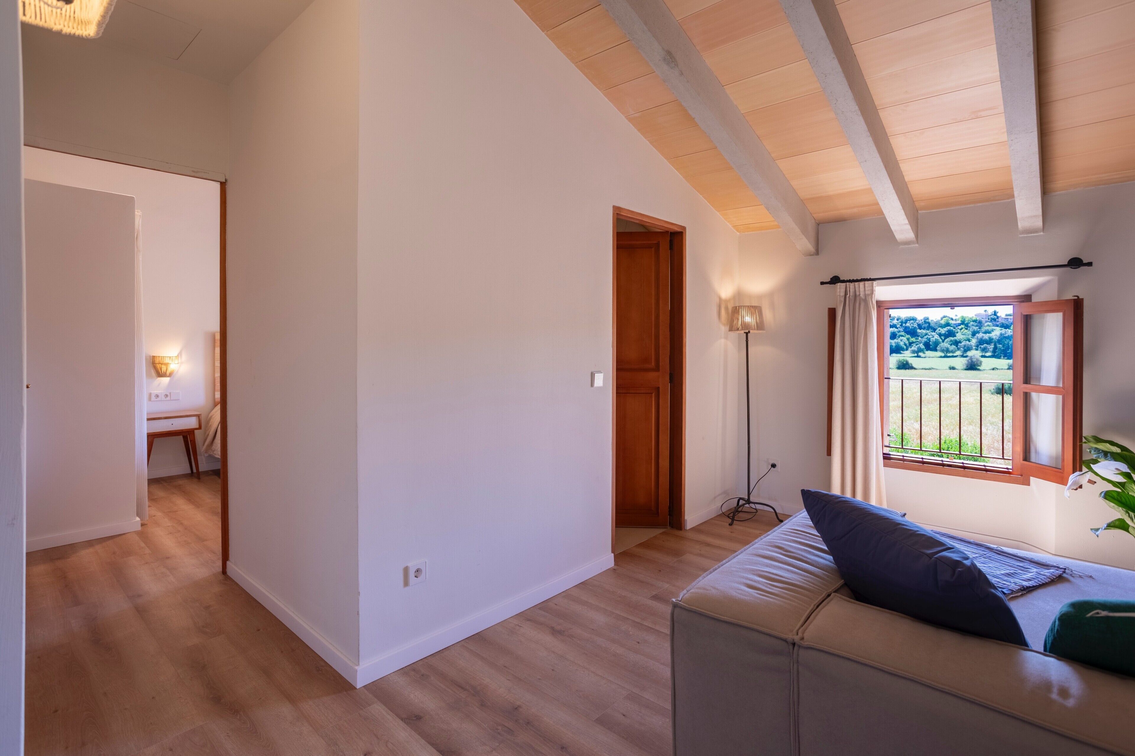 A room with a wooden floor, a sofa with cushions, a window with a view of greenery, and a door leading to another room.