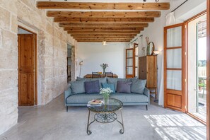 A living room with a stone wall, wooden ceiling beams, a sofa, a glass coffee table, and a view through a door. - Fantastic villa with private pool in Santa Margalida (Santa Margalida)