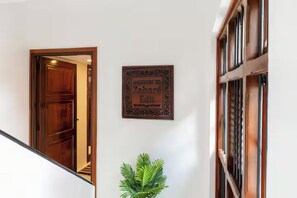 Une porte en bois donnant sur une pièce, une pancarte d’accueil encadrée et une plante en pot.