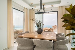 A modern dining area with a wooden table and six chairs, a vase with plants, and a view of the sea through large windows. - Moana 4 - Luxury Home with Private Pool (Benalmádena)