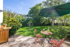 A patio with a stone floor, a wooden table, two red chairs, a green umbrella, and a small garden area.