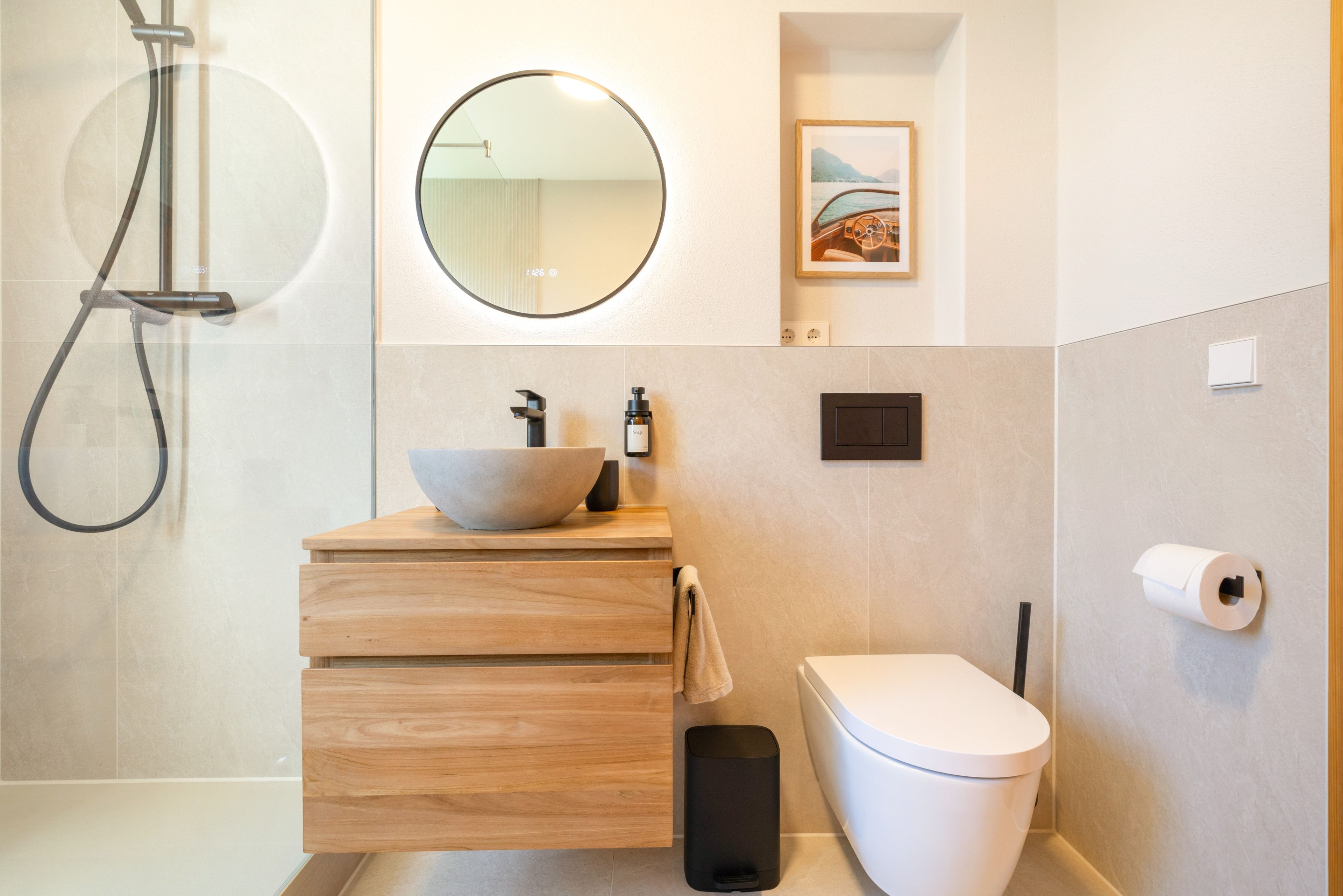 Ein modernes Badezimmer mit einer Duschkabine aus Glas, einem runden Spiegel, einer Holzwaschtischauflage mit einer Spüle, einer Toilette und einem gerahmten Bild an der Wand.