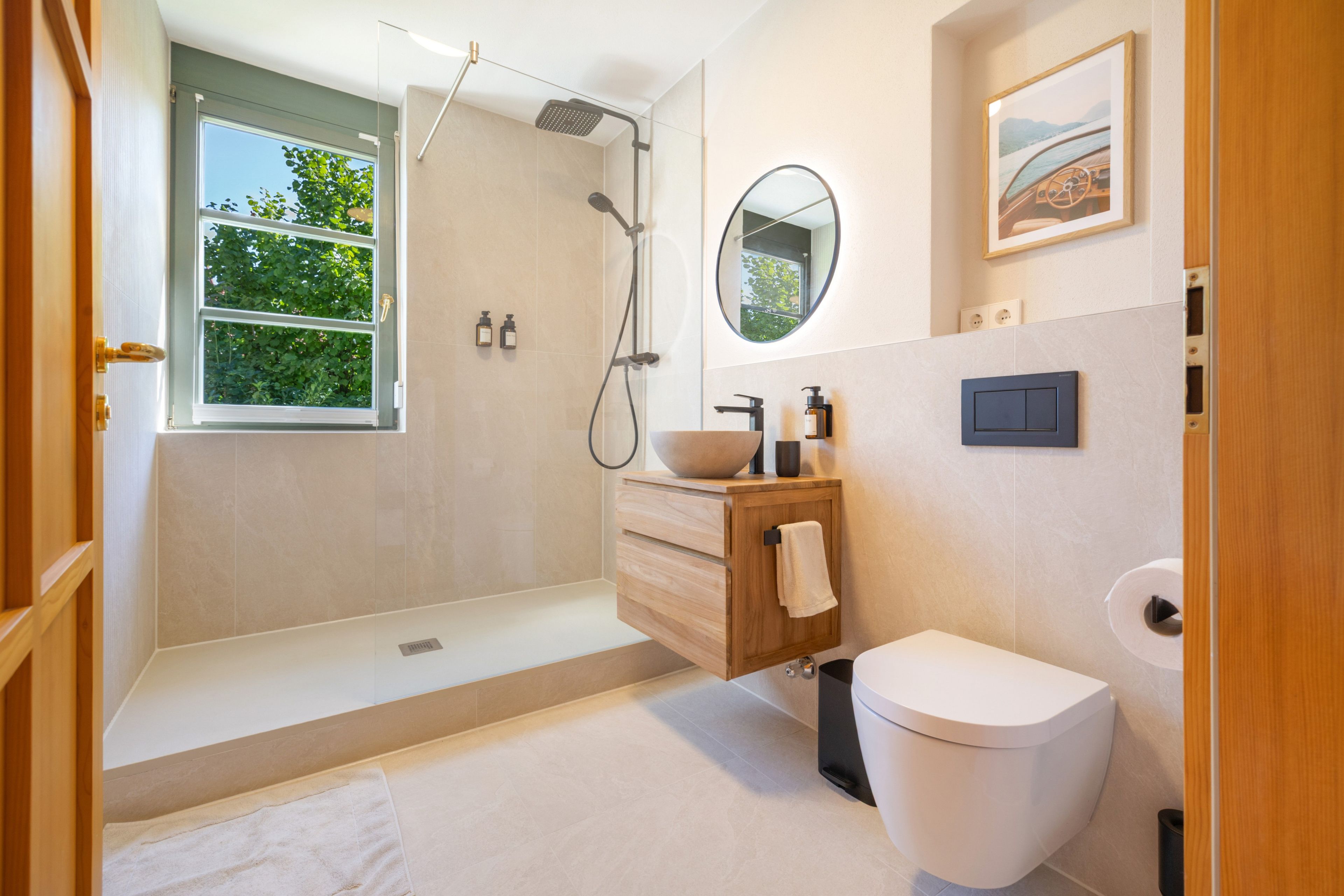 Une salle de bain moderne dotée d’une douche à l’italienne, d’un meuble-lavabo en bois, de toilettes et d’une fenêtre laissant entrer la lumière naturelle.