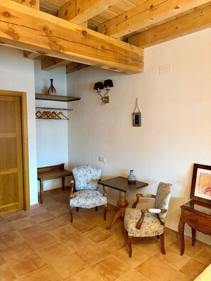 Quarto com vigas de madeira, um banco, duas cadeiras, uma mesa e uma luminária fixada na parede.