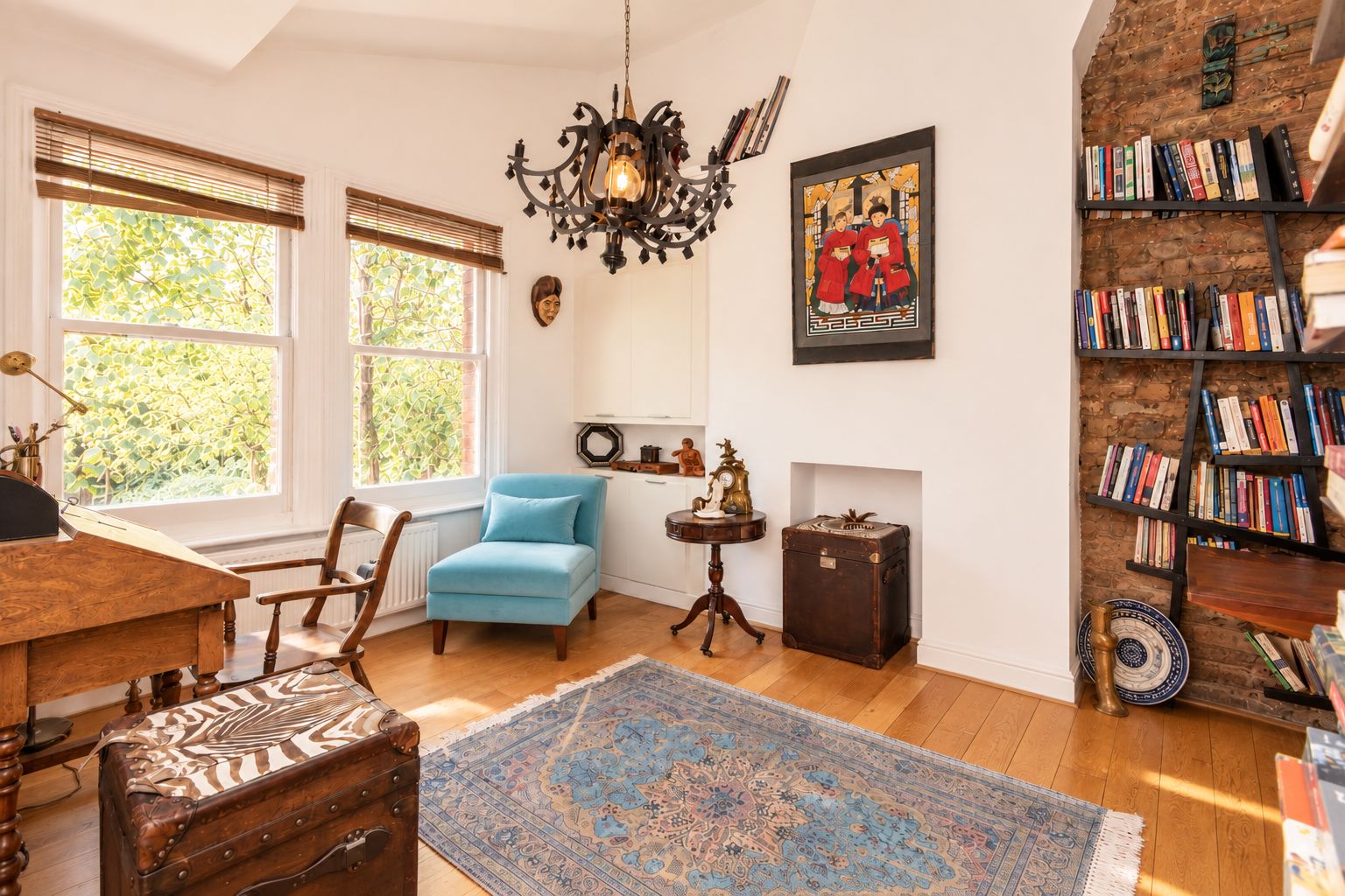 A room with a blue armchair, wooden floor, a patterned rug, a bookshelf, and a wall-mounted painting.