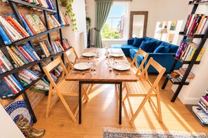 A dining area with a wooden table set for four, surrounded by bookshelves filled with books, a blue sofa, and a window with curtains. - 2-Bed Chelsea Flat | Balcony & River View (London)